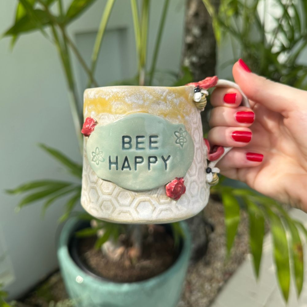 Hand holding a ceramic mug with 'BEE HAPPY' text, surrounded by plants.